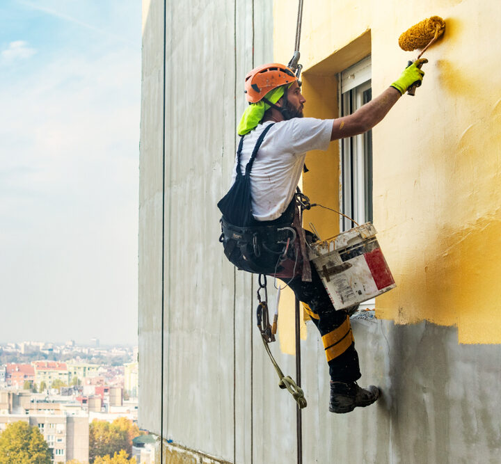 Painter painting exterior walls of commercial building hanging from rope supports with paint roller and bucvket of paint - Quillen Construction Group