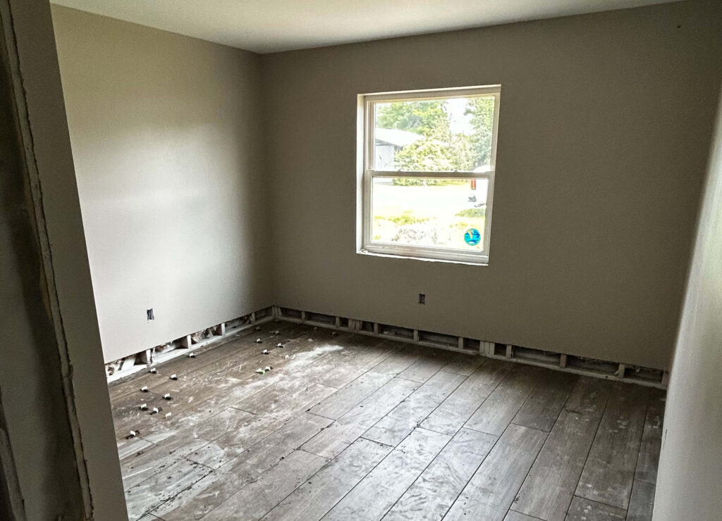 An empty room with beige walls, a single window, and a bare floor with debris—ideal for water damage restoration. The baseboards are missing, exposing the wall framing as natural light enters through the window.
