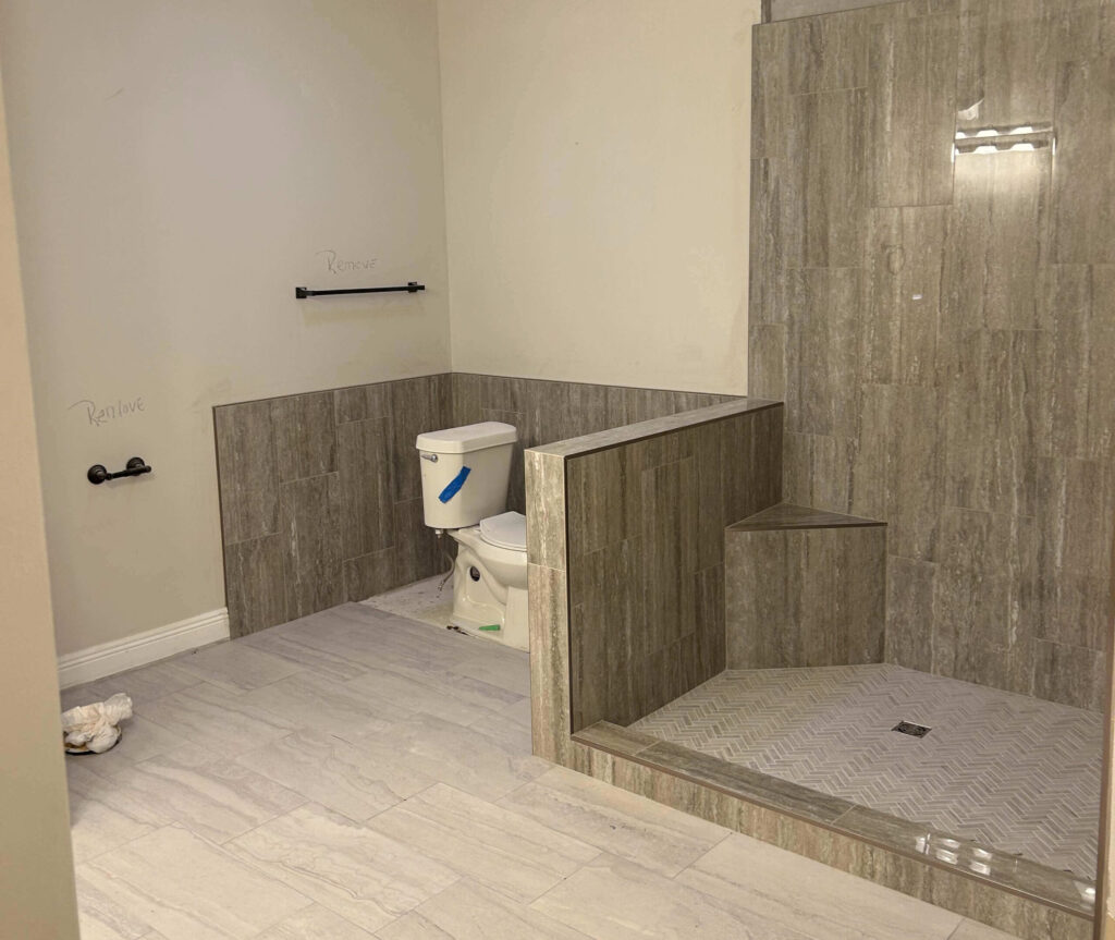 A bathroom with beige tile flooring and walls, featuring a toilet next to a walk-in shower with a built-in bench. Signs of water damage are visible near the toilet paper holder and towel bar, where “Replace” is written on the wall.