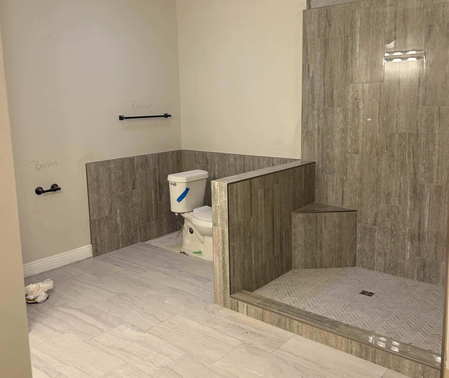 A bathroom with beige tile flooring and walls, featuring a toilet next to a walk-in shower with a built-in bench. Signs of water damage are visible near the toilet paper holder and towel bar, where “Replace” is written on the wall.