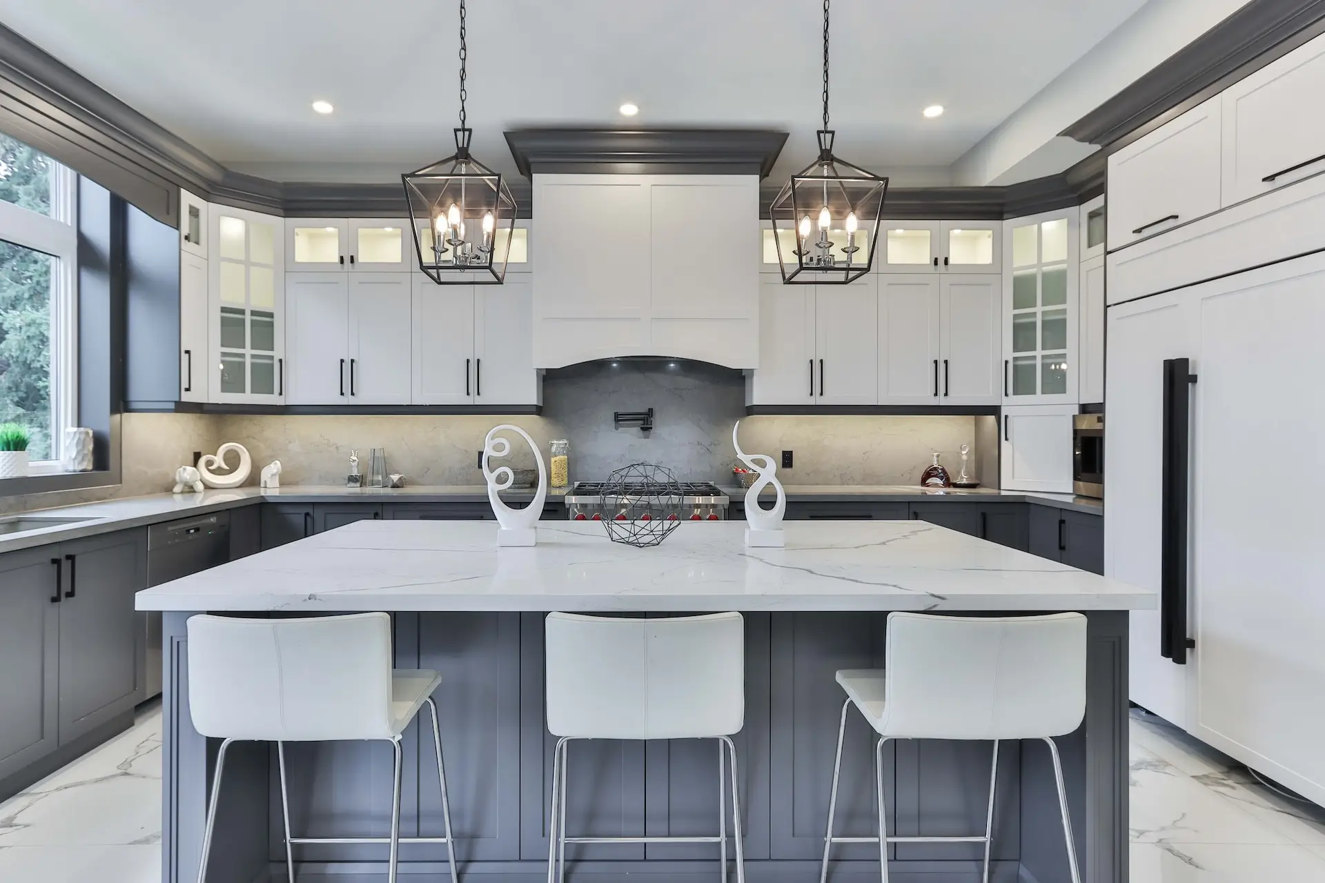 Kitchen Remodel New Orleans - An image of a modern kitchen with a custom white fridge with black handles, custom island in an off-grey, white leather upholstered stools, custom concrete backsplash, custom cabinets with glass windows and interior lighting.