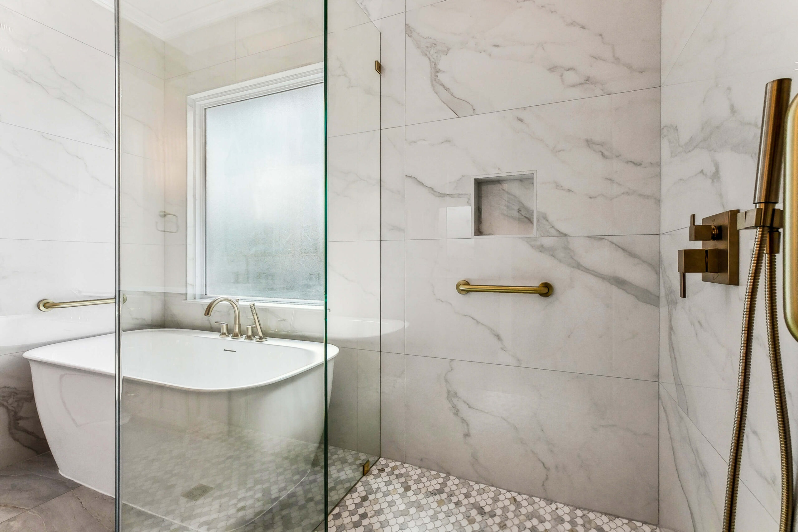 Modern bathroom with a white freestanding bathtub by a frosted window, marble-patterned walls, a glass shower enclosure, gold fixtures, and a built-in shower niche.