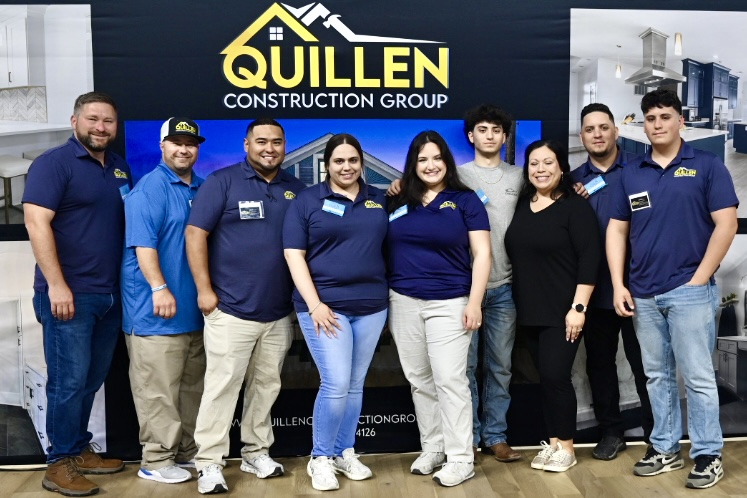 A group of nine people, wearing blue shirts with logos, stand together smiling in front of a "Quillen Construction Group" banner at an indoor event.