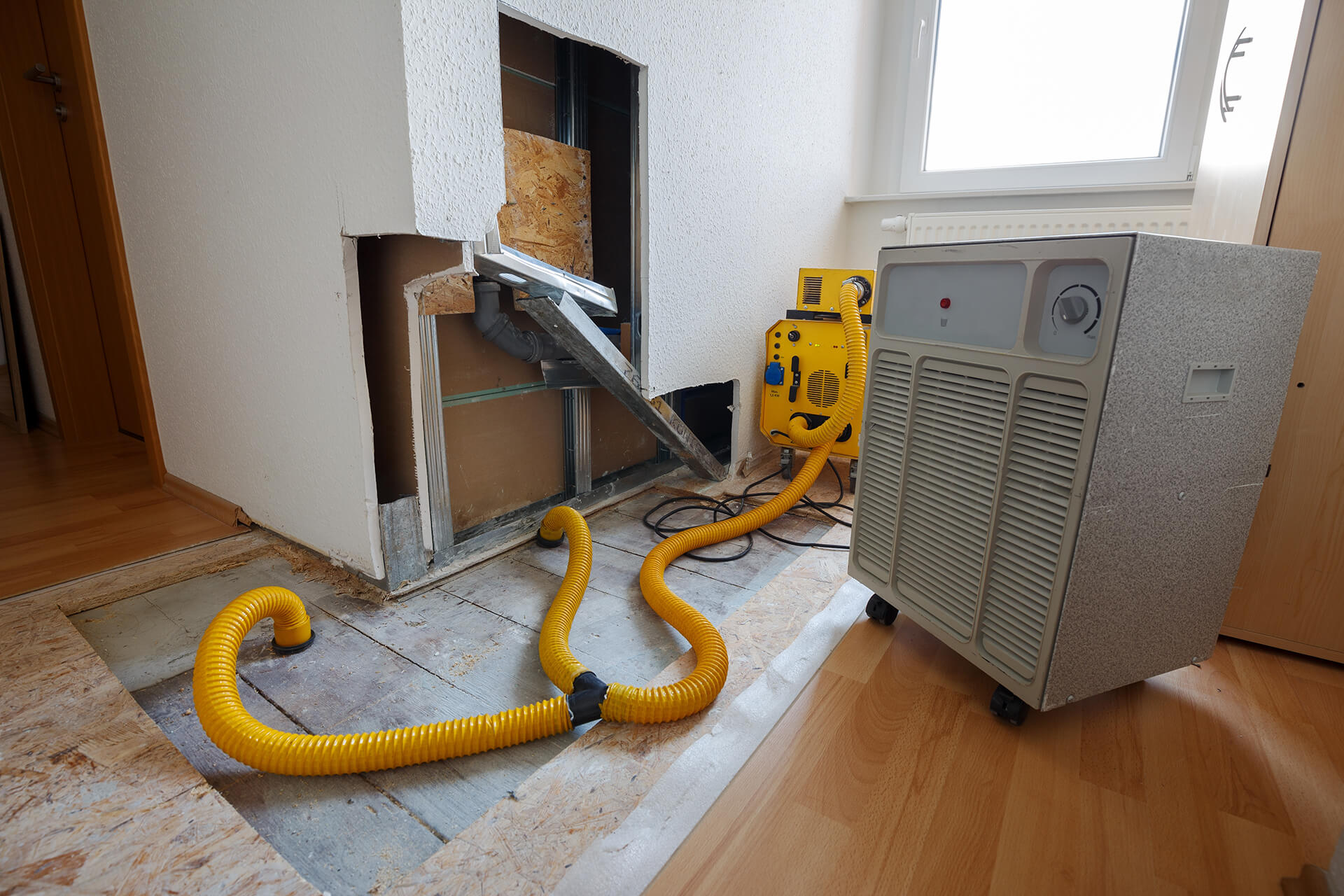 A room with a section of wall and floor removed shows exposed pipes. Two yellow hoses connect a yellow machine and a large silver dehumidifier, highlighting Damage Restoration New Orleans is underway to repair water damage and dry the area.