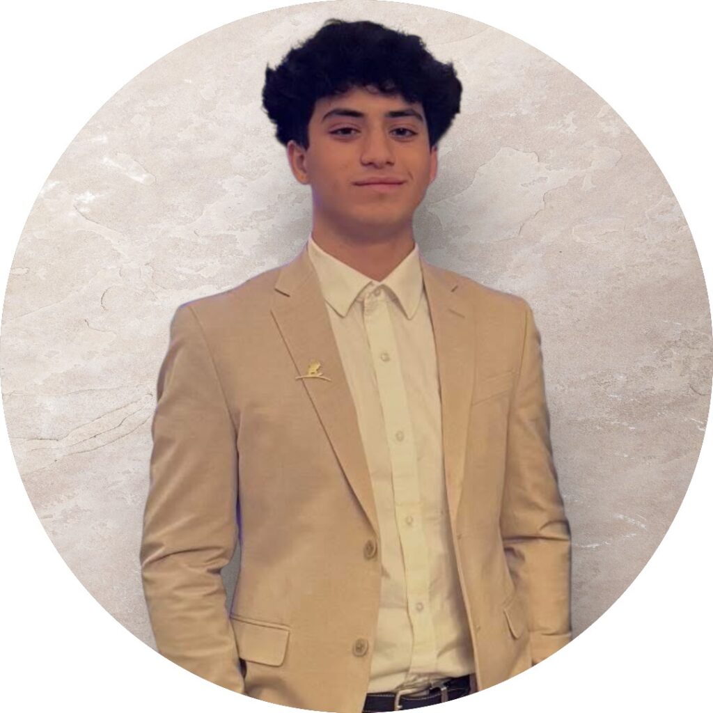 A young person wearing a tan suit jacket, white dress shirt, and no tie stands in front of a light-colored, textured background, facing the camera with a slight smile.