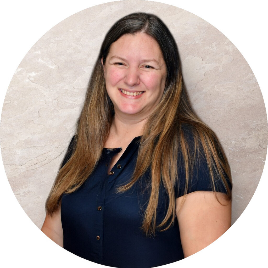 A woman with long brown hair wearing a short-sleeved dark blue button-up shirt stands smiling in front of a light beige textured background.