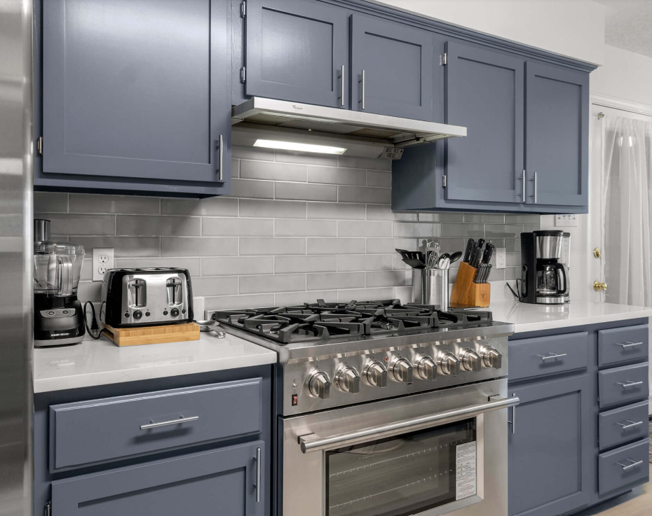 A modern kitchen features blue cabinets, a stainless steel gas stove with oven, a gray subway tile backsplash, and countertop appliances including a toaster, blender, knife block, and coffee maker.