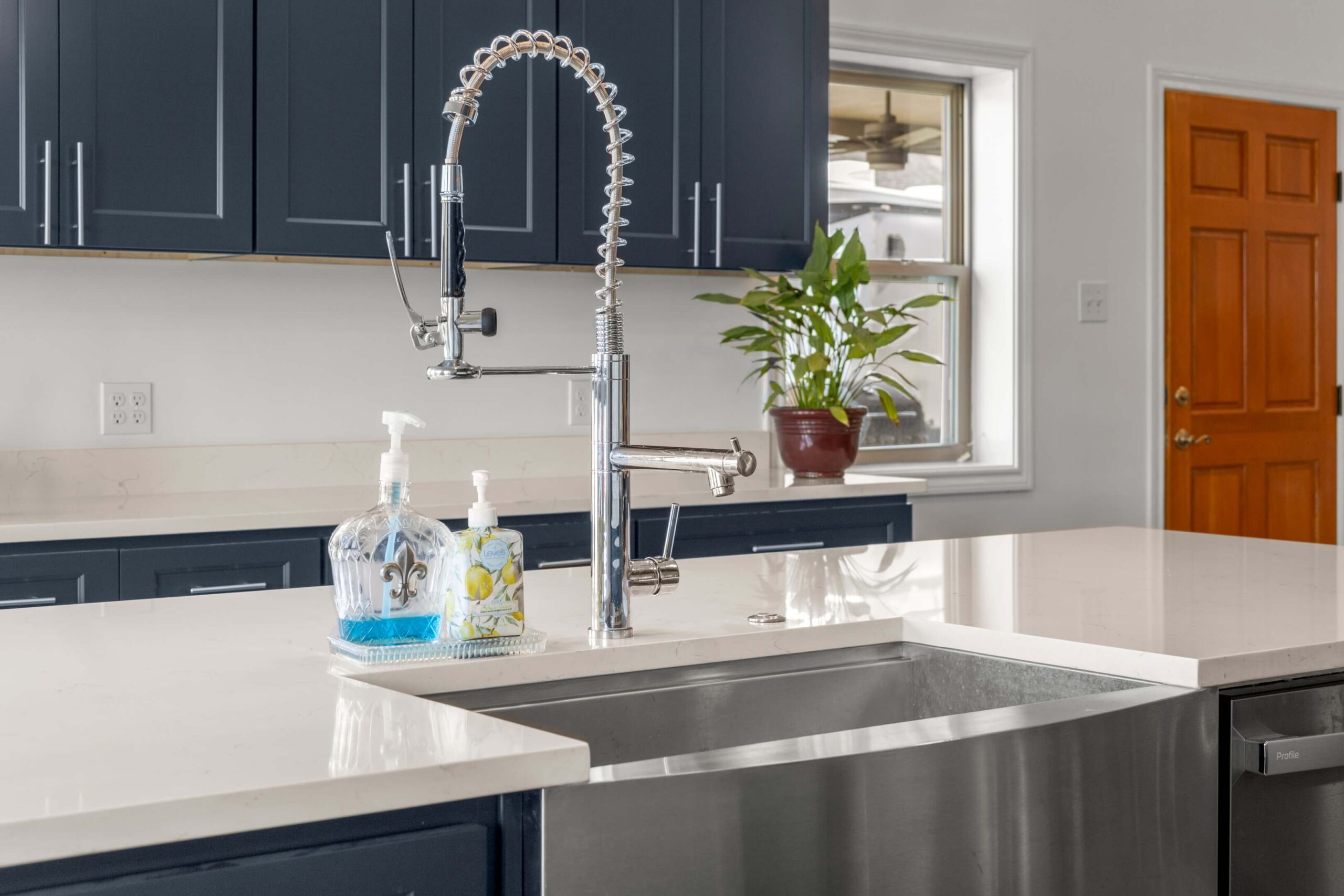 A modern Covington kitchen countertop with a deep stainless steel sink, a tall spring pull-down faucet, blue lower cabinets, soap dispensers, a potted plant by the window, and a wooden door shows expert kitchen remodeling.