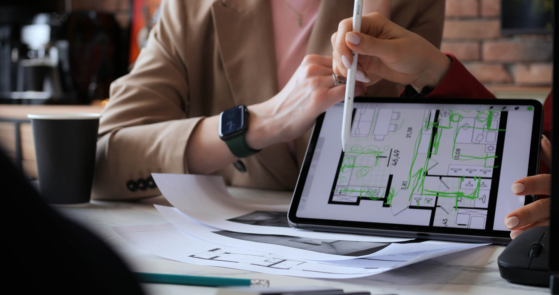 Two people review Elmwood kitchen remodeling floor plans displayed on a tablet, with one person pointing at the screen using a pen. Papers and architectural drawings are spread out on the table in front of them.