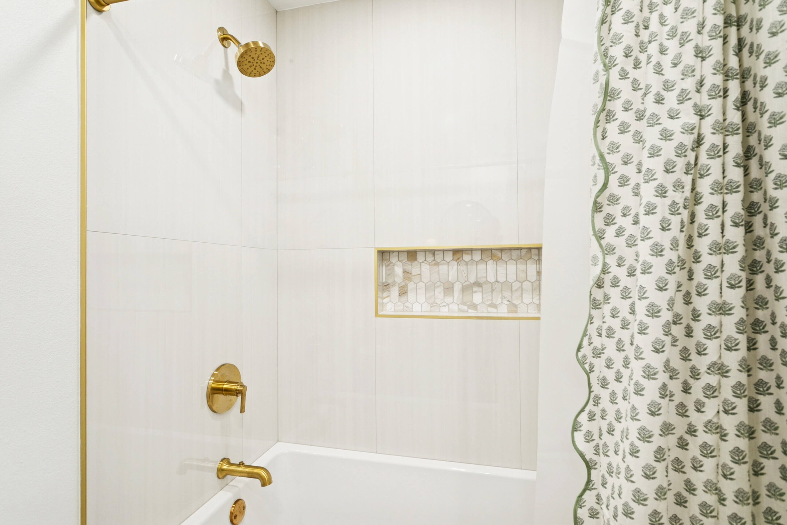 A bathroom shower in Gretna, LA, features white tile walls, gold fixtures, a built-in shower niche with small decorative tiles, and a white shower curtain with a green leaf pattern—perfect inspiration for your next bathroom remodeling project.