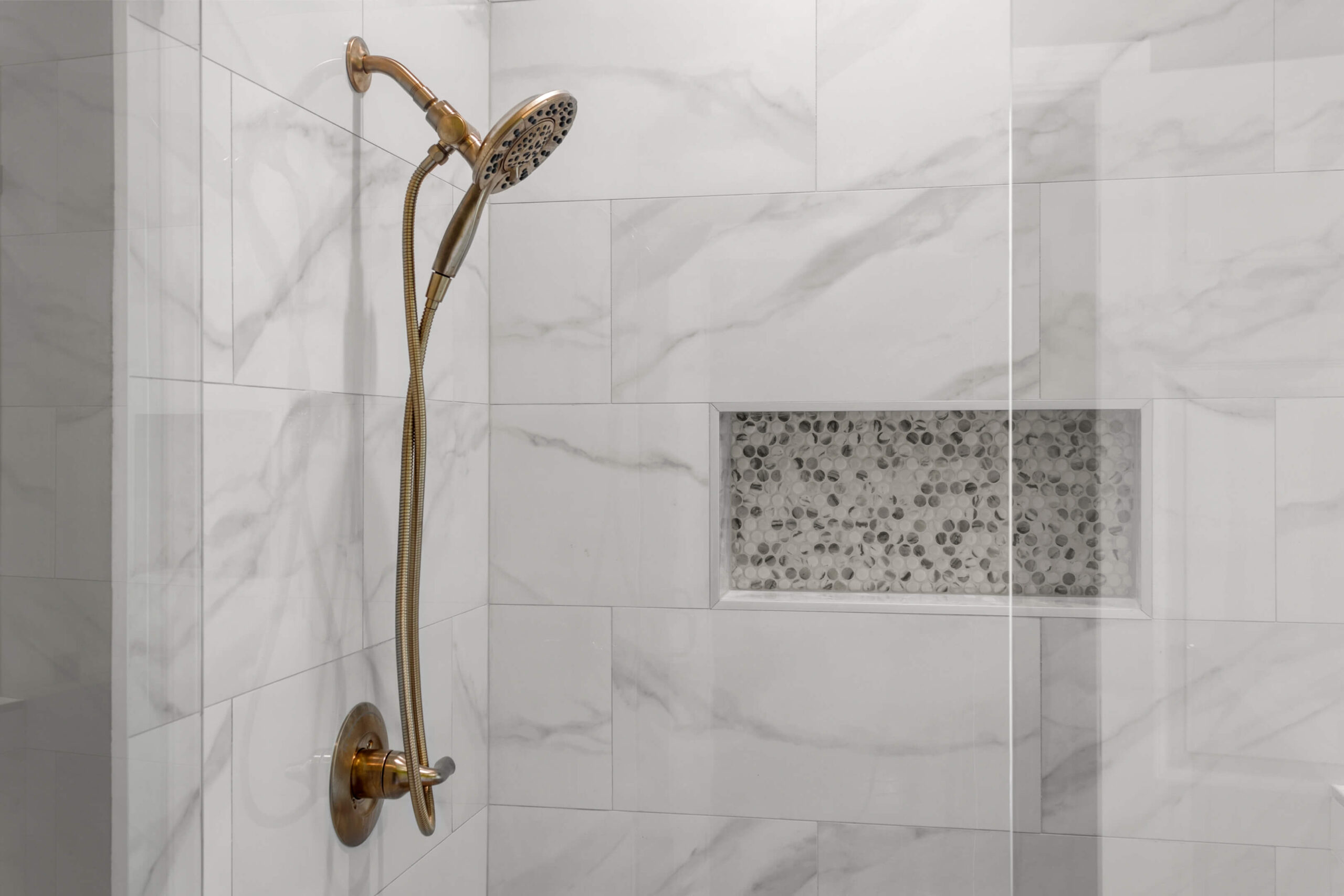 A shower with a brushed gold handheld showerhead and hose is mounted on a wall with white and gray marble tiles—perfect for your next Bathroom Remodeling project in Belle Chasse, LA. There’s also a built-in niche with small round gray and white mosaic tiles.