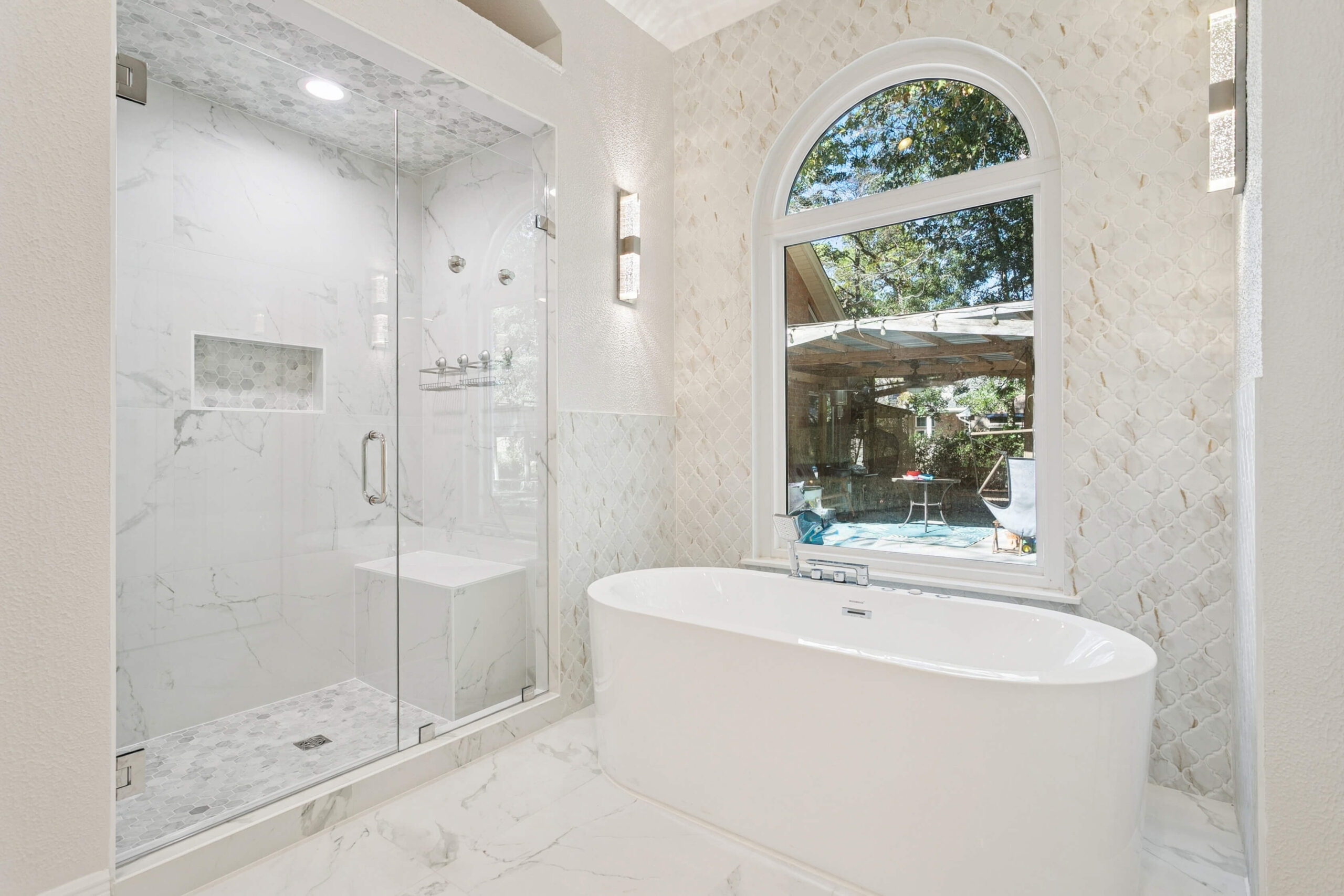 A modern LA bathroom with a glass-enclosed shower, white freestanding bathtub, marble floors, and a large arched window overlooking trees and patio furniture—a stunning Bathroom Remodeling project in Terrytown.