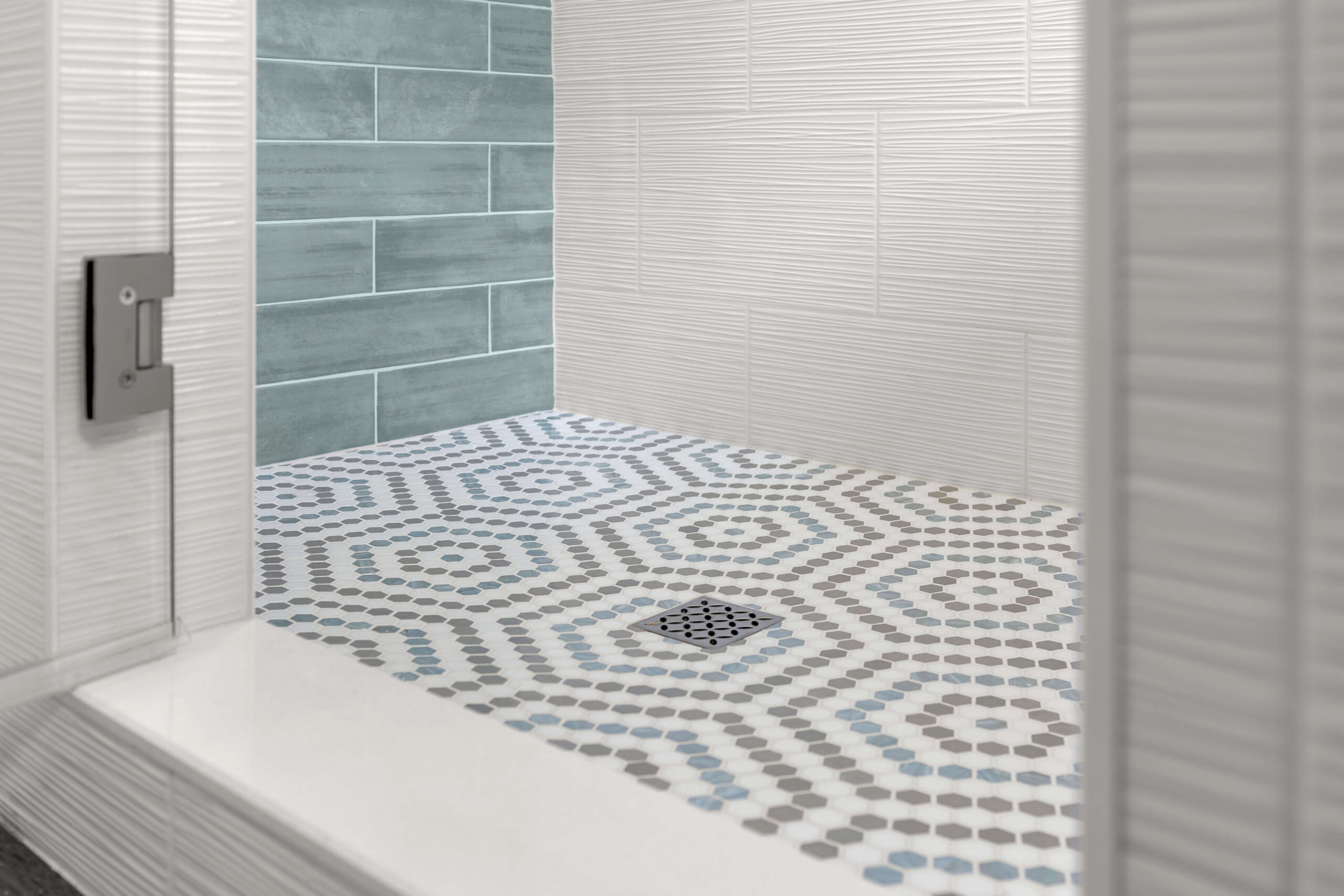 A modern Lakeview bathroom remodeling features a shower with small hexagonal tiles in gray, white, and blue forming a geometric pattern. The walls showcase textured white and rectangular blue tiles, with a square drain centered on the floor.