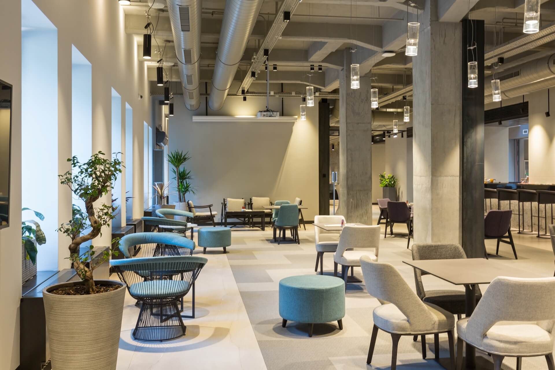 A modern, spacious lounge area—ideal for commercial construction in Jefferson, Louisiana—features various chairs, tables, ottomans, potted plants, exposed ducts, hanging lights, and large windows that flood the space with natural light.
