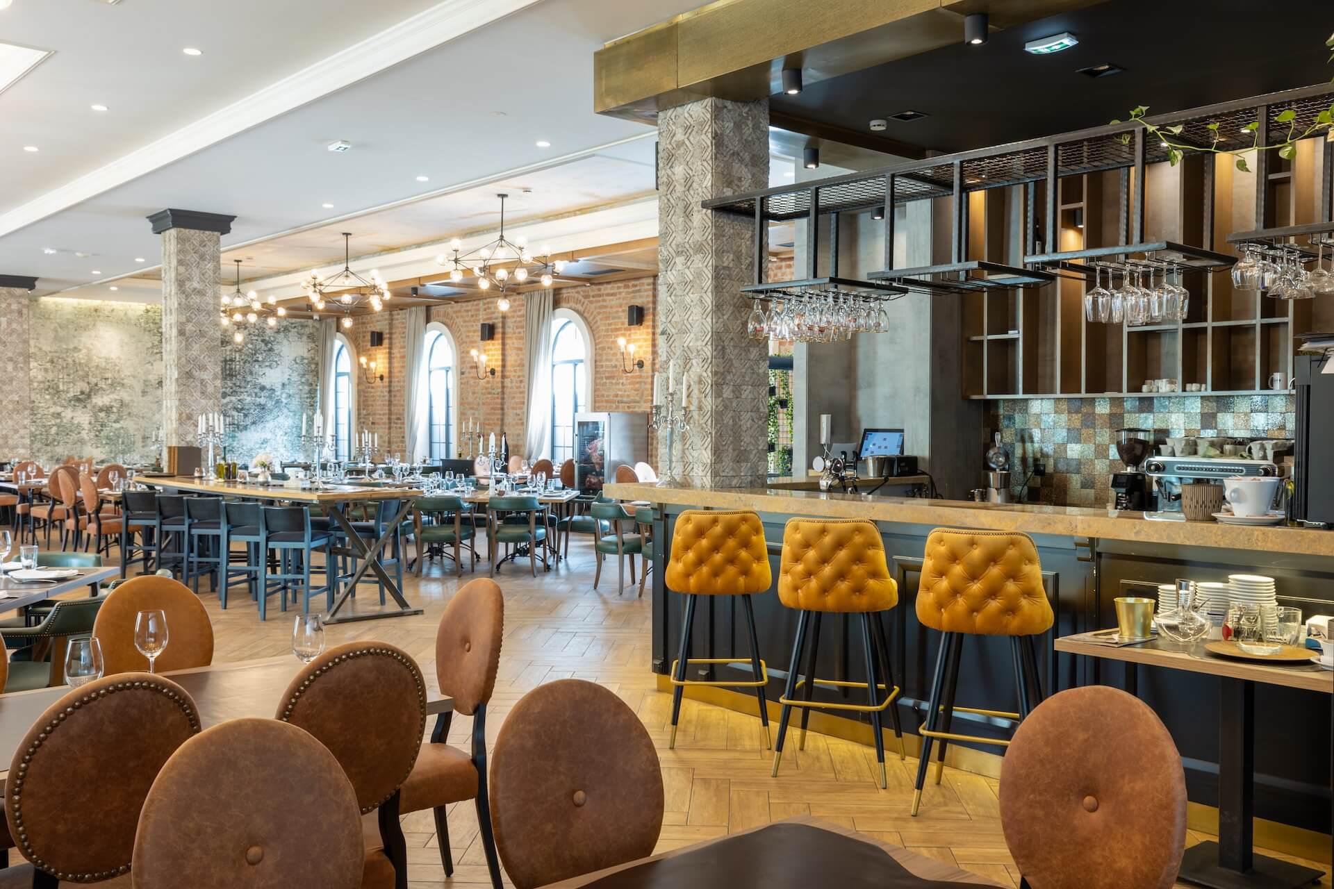 A spacious, modern restaurant in Violet, Louisiana, featuring brown leather chairs, wooden tables, a bar area with yellow barstools, hanging glasses, and exposed brick walls with large arched windows—showcasing expert commercial construction.