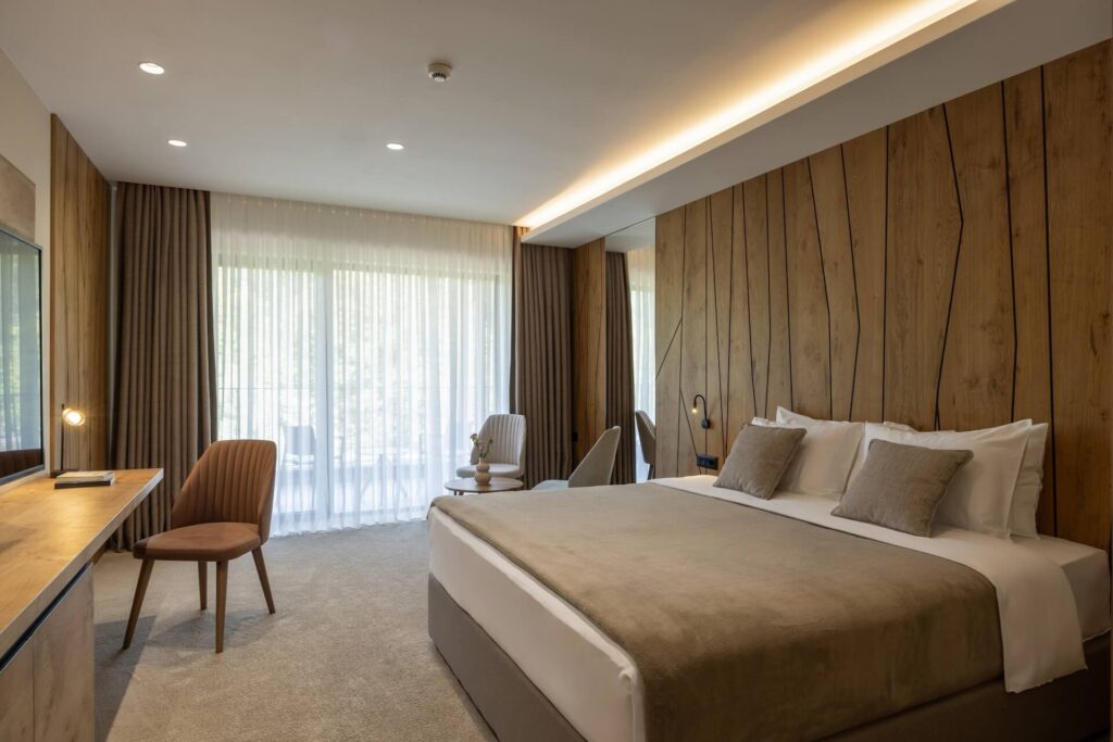 A modern hotel room in Lakeview, New Orleans, featuring a large bed, neutral bedding, and wooden accent wall—designed with quality commercial construction. Enjoy a desk workspace and cozy table for two by the window with floor-to-ceiling curtains.