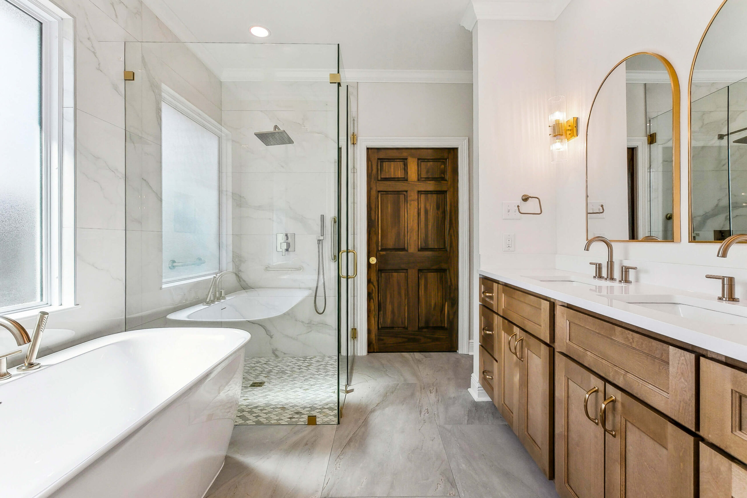 A modern bathroom by a leading construction company in New Orleans features a freestanding bathtub, glass-enclosed shower, double sink vanity with wooden cabinets, large mirrors, gold fixtures, gray tile floor, and a wooden door at the back.