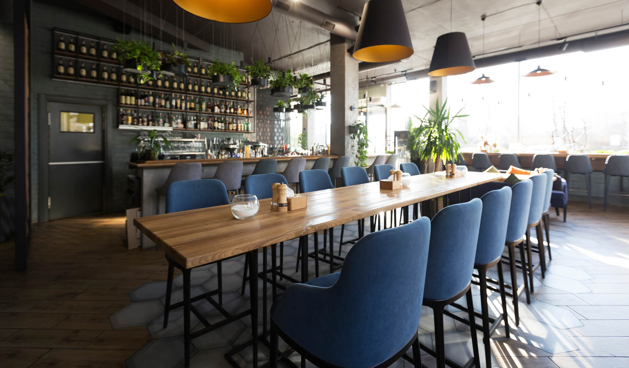 A modern restaurant interior, crafted by a leading construction company in Covington, LA, features a long wooden table with blue upholstered chairs, hanging plants, striking pendant lights, and a backlit bar. Sunlight pours in through expansive windows.
