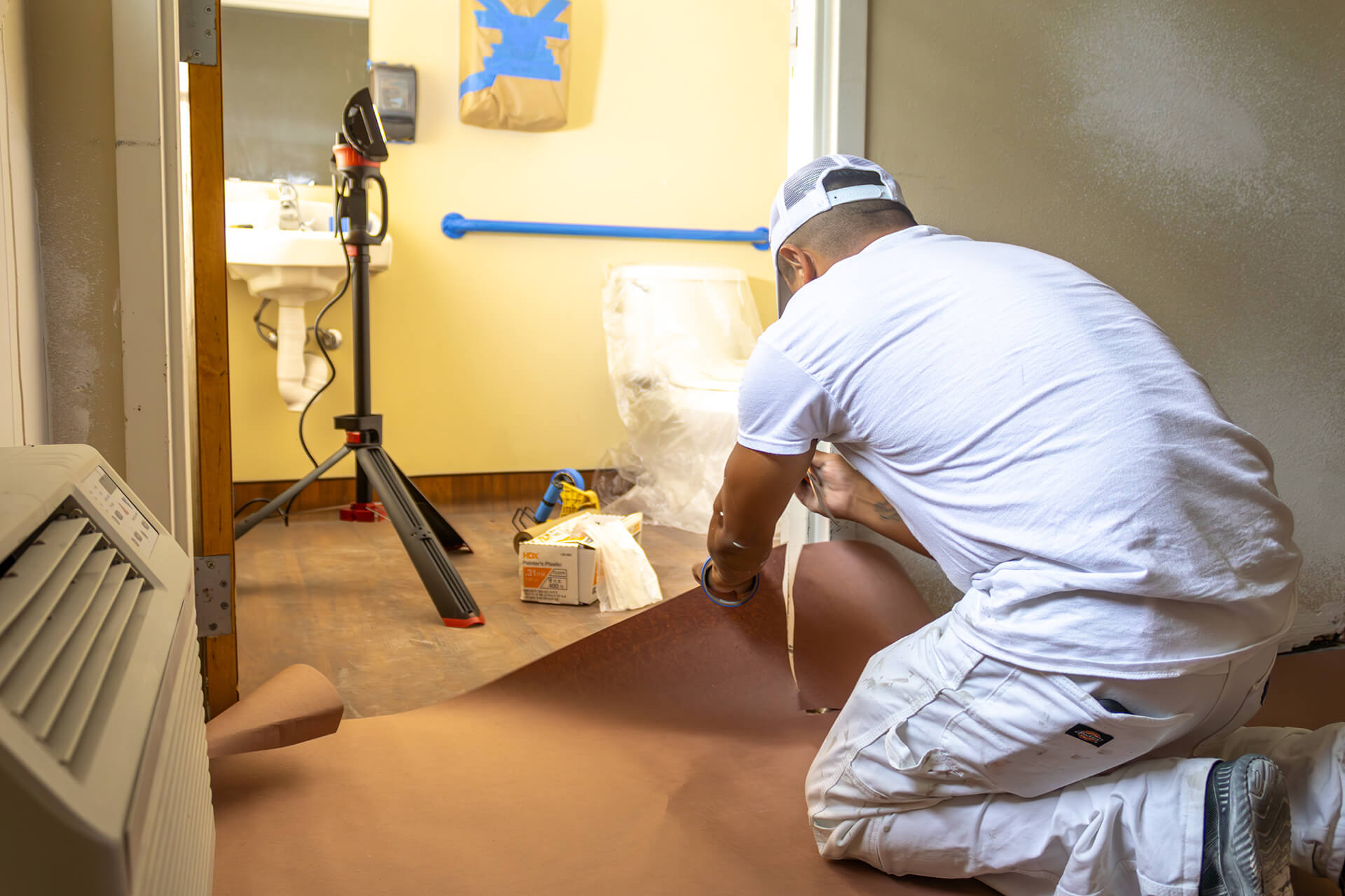 A general contractor in white work clothes covers the floor with brown protective paper in a Hammond, LA room being prepared for painting. A sink, taped surfaces, and painting tools are visible in the background.