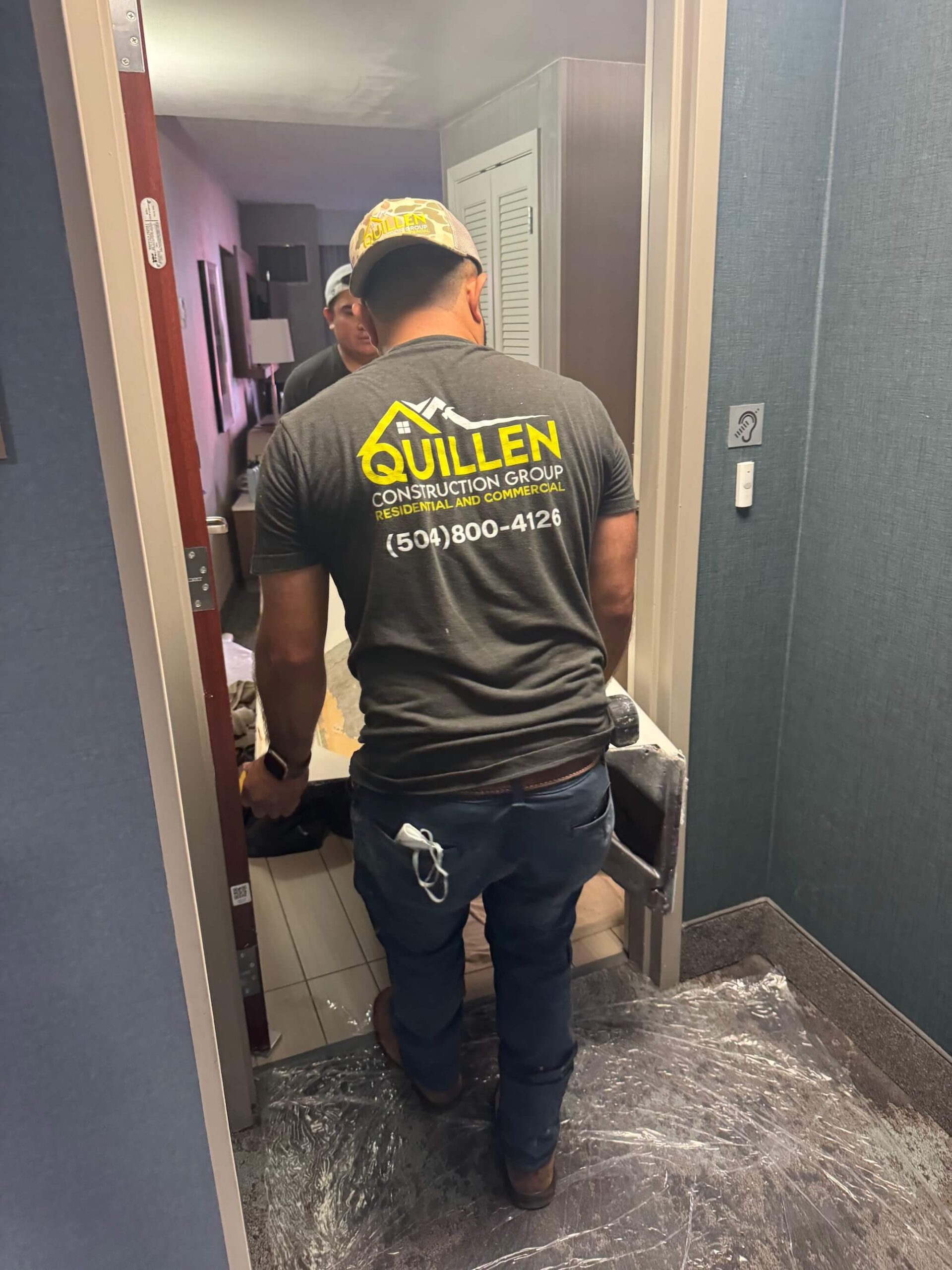 A man wearing a "Quillen Construction Group" t-shirt and cap stands in a doorway in Madisonville, LA, with another person in the background. Tools and protective floor covering suggest this general contractor is busy on site.