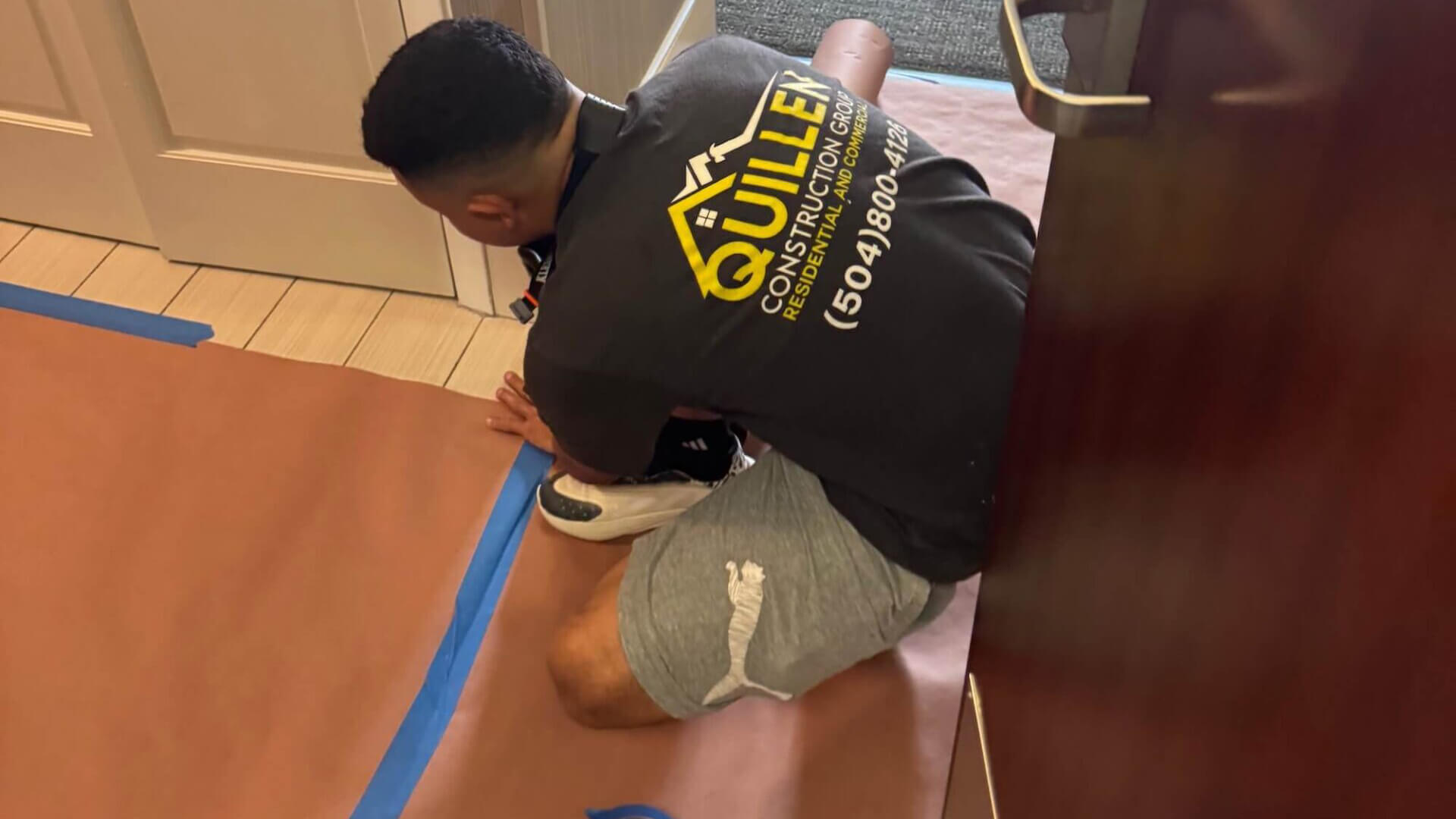 A person wearing a Quillen Construction Group t-shirt and shorts kneels on the floor, securing brown protective paper with blue tape near a doorway for a construction company Jefferson LA project.