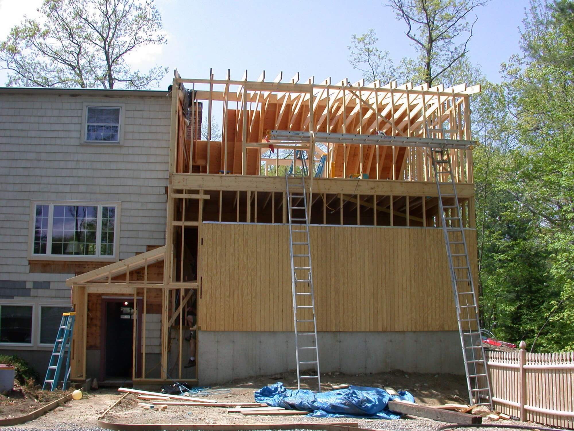 A two-story home addition in Harahan, LA under construction features exposed wooden framing, plywood sheets, two ladders leaning against the structure, and construction materials scattered on the ground.