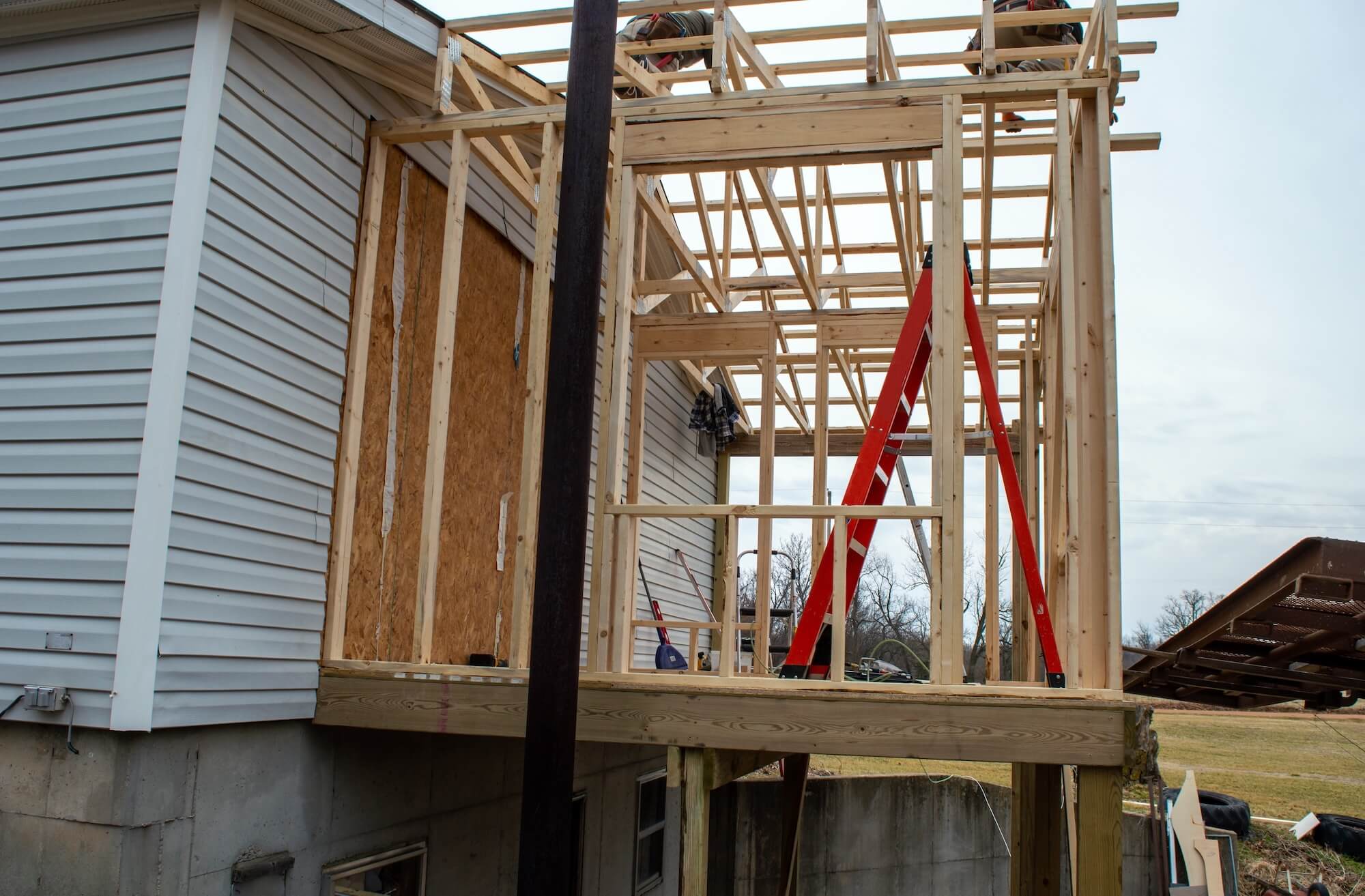 A wooden home addition is under construction in Marrero, LA, with exposed framing, a red ladder in the center, and an unfinished wall attached to a white-sided house. Tools and building materials are visible in the background.