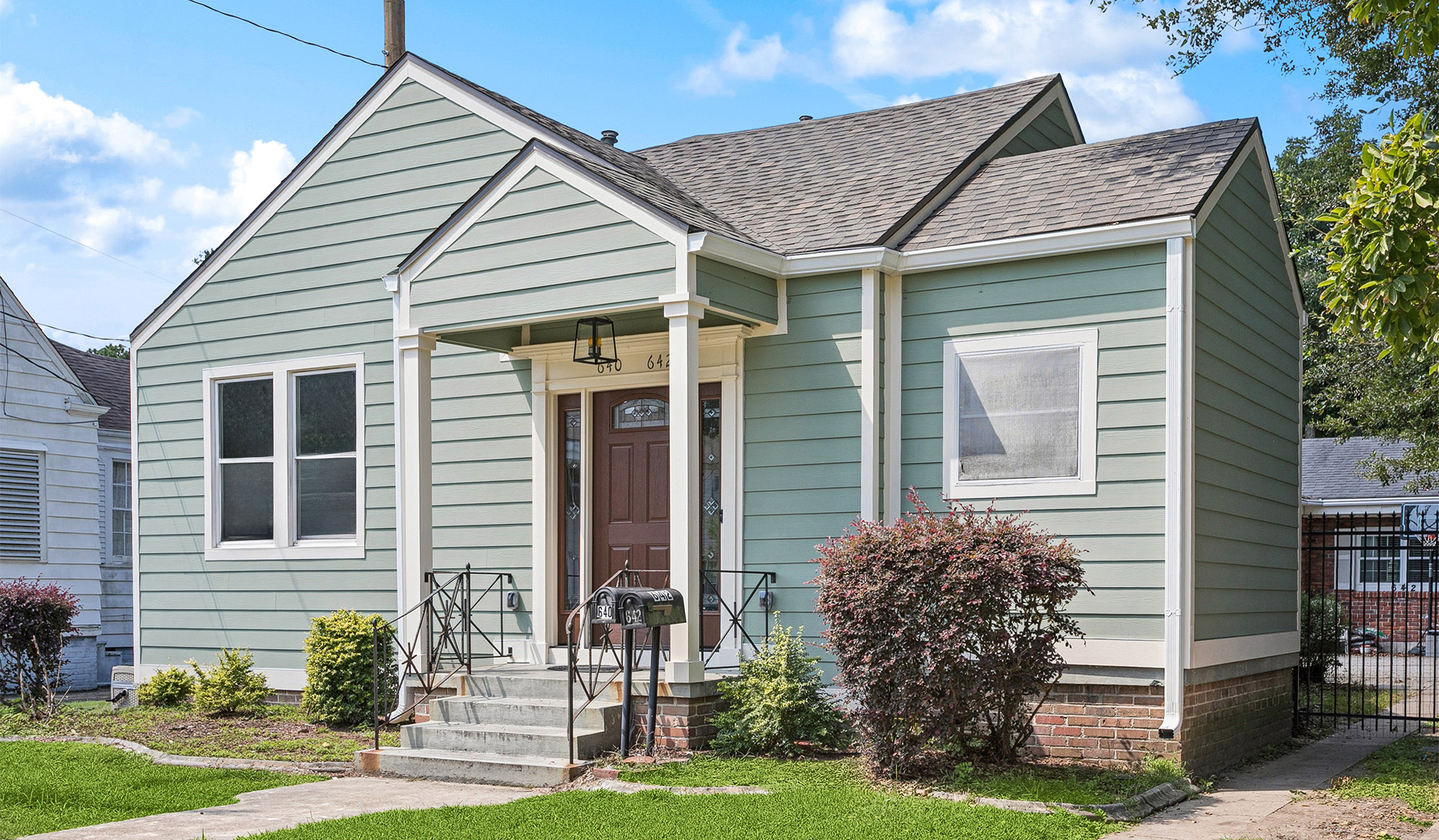A small, light green house with white trim, a brown front door, a covered entryway with metal railings—expertly built by a top construction company in Arabi, LA—features two large front bushes, a short staircase, and a green lawn in front.