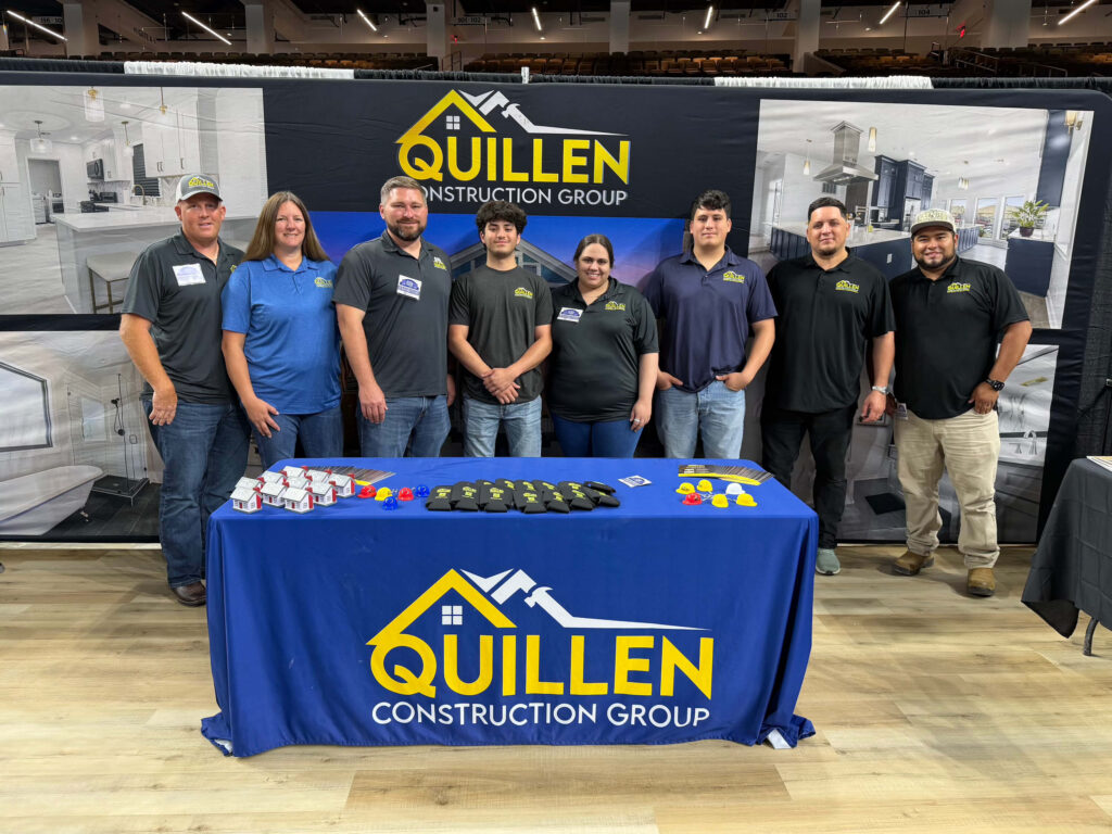 Nine people stand behind a display table with promotional items and a blue tablecloth featuring the Quillen Construction Group logo. A branded backdrop with construction images is behind them in a large indoor setting.