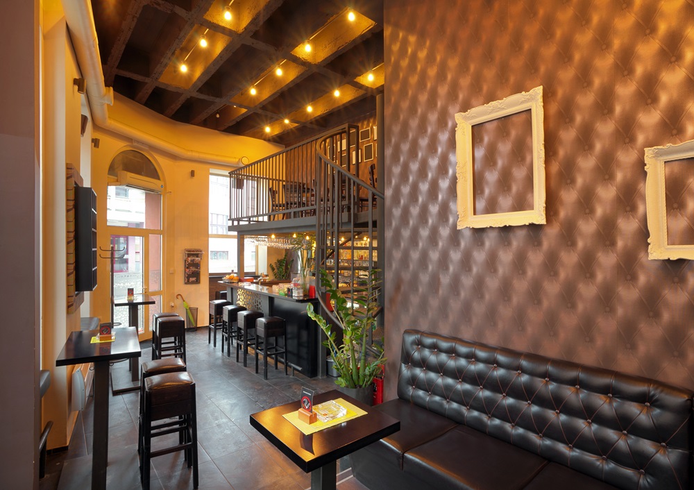 A modern cafe interior with brown quilted walls, empty picture frames, black leather seating, high ceilings with exposed beams, small tables—perfect for small dining area ideas—a mezzanine level, and warm ambient lighting.