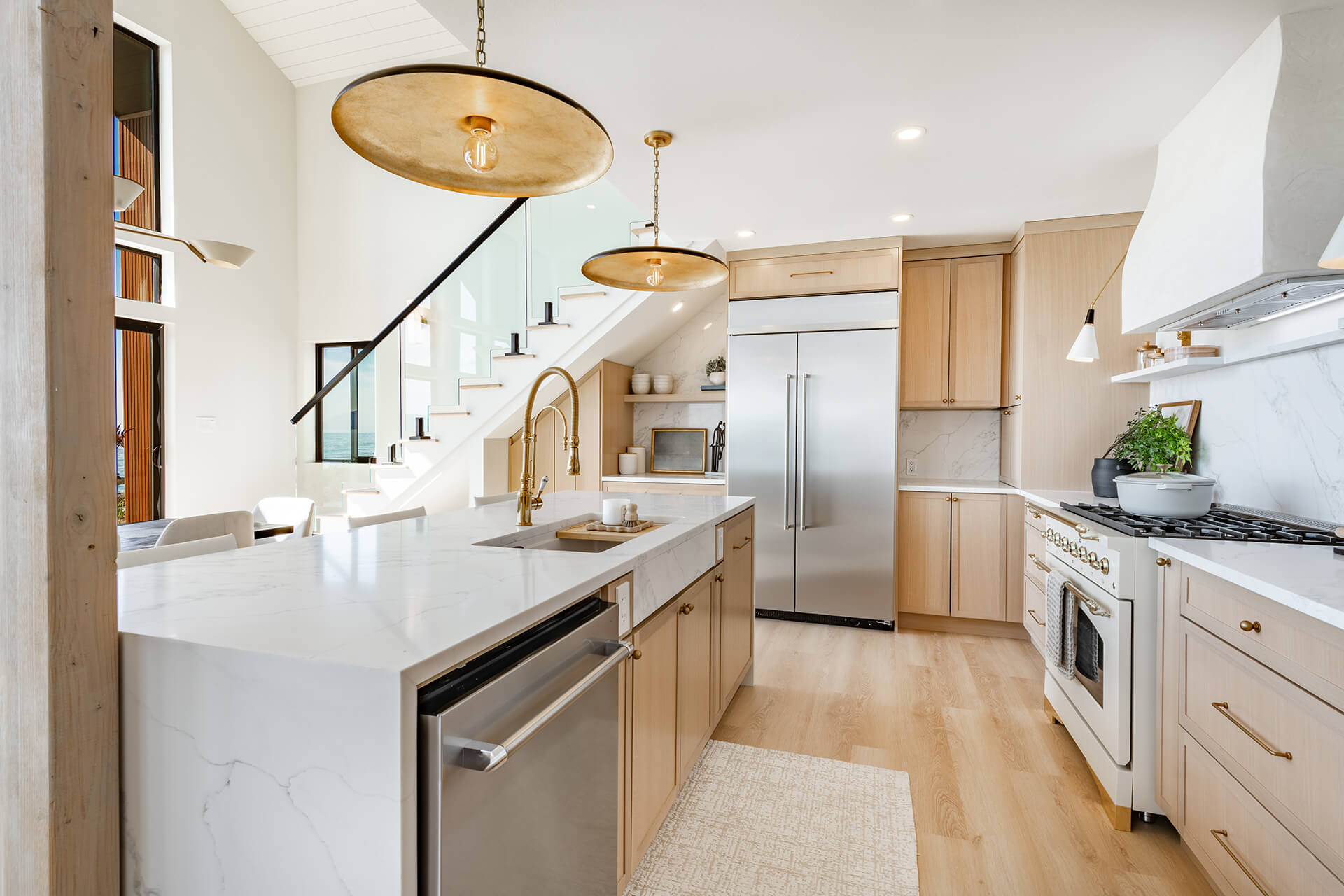 Modern kitchen designed by a construction company in Avondale, LA, featuring light wood cabinets, white marble countertops, stainless steel appliances, a large refrigerator, gold faucet, pendant lights, and a staircase with a glass railing in the background.