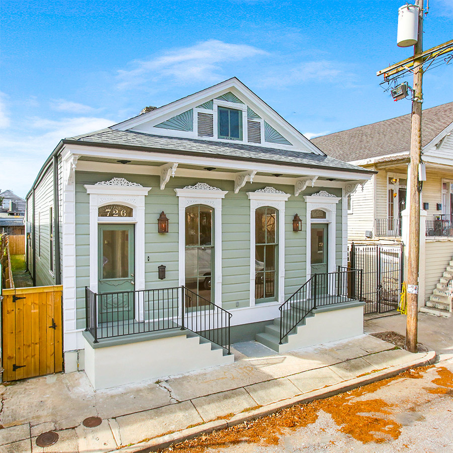 A light green single-story house in Chalmette with white trim, tall windows, decorative gables, and two front entrances. A black metal railing lines both sets of steps, and a wooden gate is on the left side. A power pole is in front.