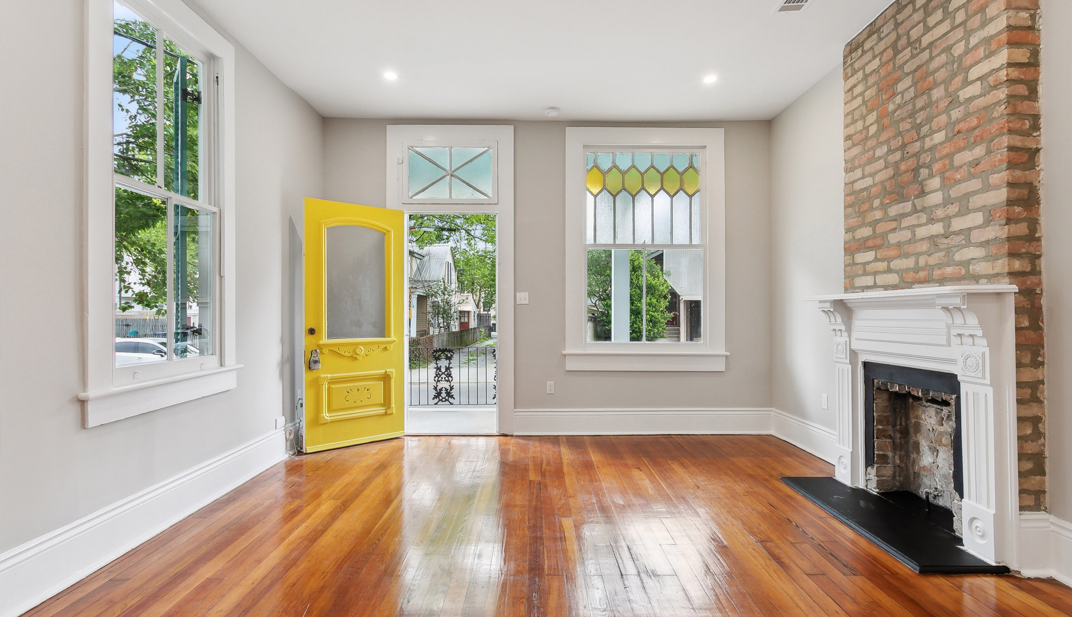 A bright living room by Violet LA with polished wood floors, a yellow front door, exposed brick fireplace, and large windows—one featuring stained glass—that beautifully showcase expert home renovations and natural light.