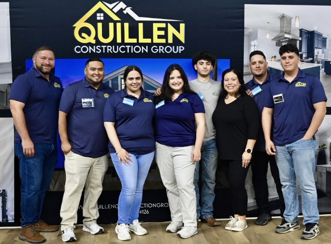 Seven people stand together in front of a Quillen Construction Group backdrop in Woodmere, LA. Most are wearing matching blue company shirts as they smile for a group photo, showcasing their Whole Home Renovations team spirit indoors.