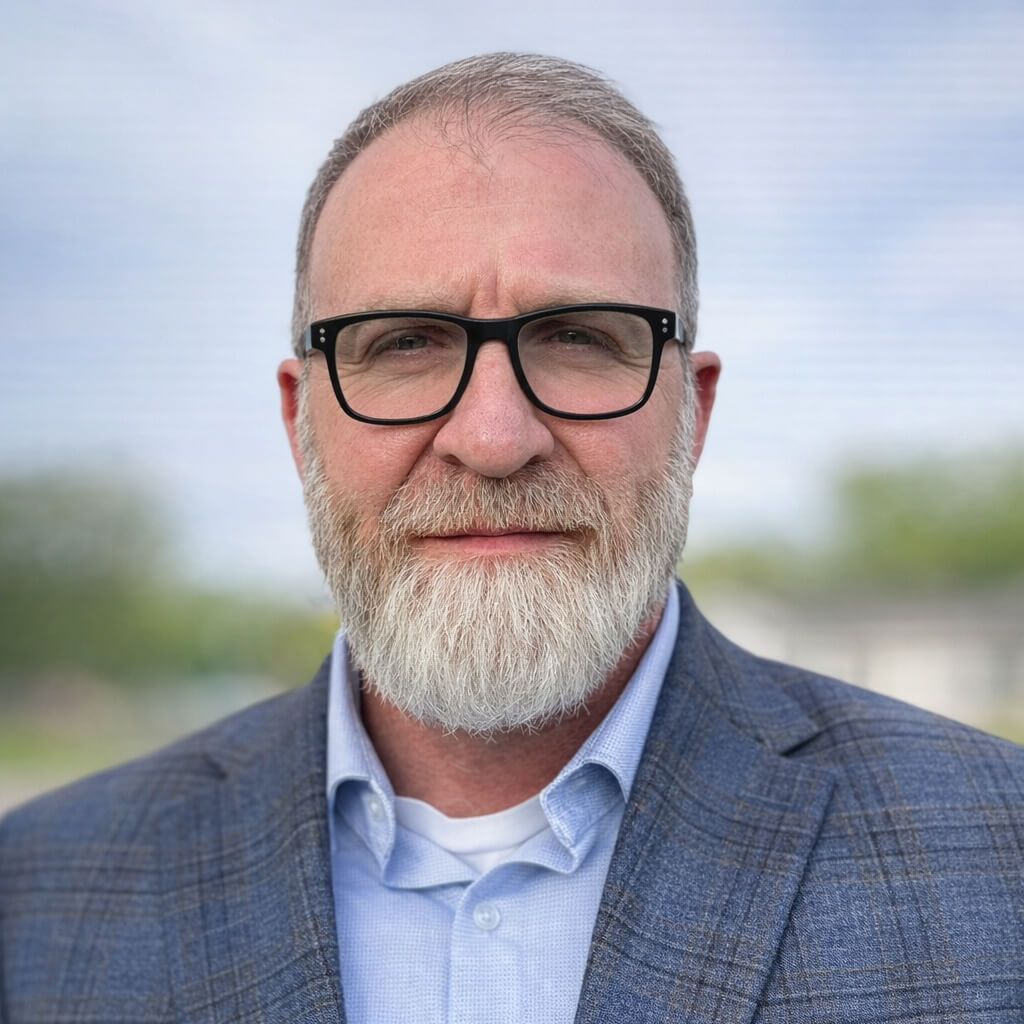 A middle-aged man with gray hair and a full beard, wearing black glasses, a light blue shirt, and a blue plaid suit jacket, standing outdoors with a blurred background.